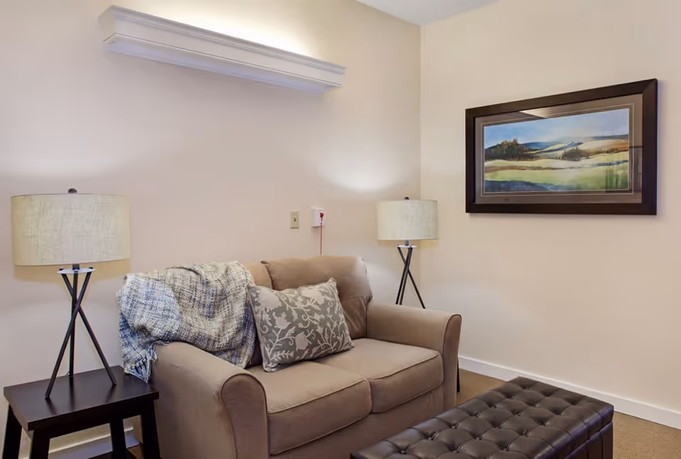 A cozy living room area with a beige loveseat adorned with a patterned throw blanket and a decorative pillow. There are two matching table lamps on either side of the loveseat, one on a small dark wooden side table. A framed landscape painting hangs on the wall above the loveseat, and a dark tufted ottoman is placed in front of it.