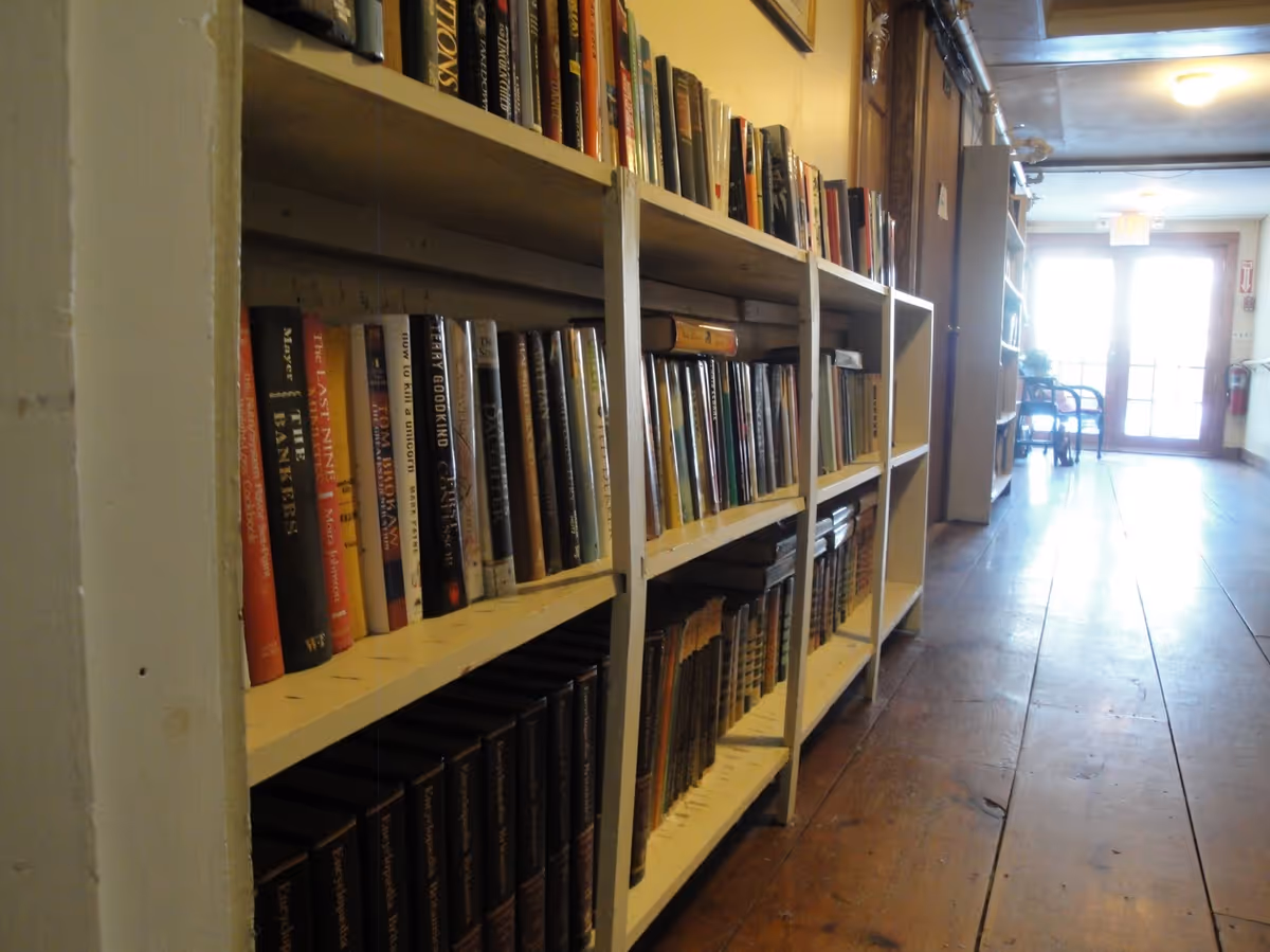 A hallway with wooden flooring and bookshelves filled with books on the left side. At the end of the hallway, there are glass double doors letting in natural light, with a chair and some plants visible near the doors.