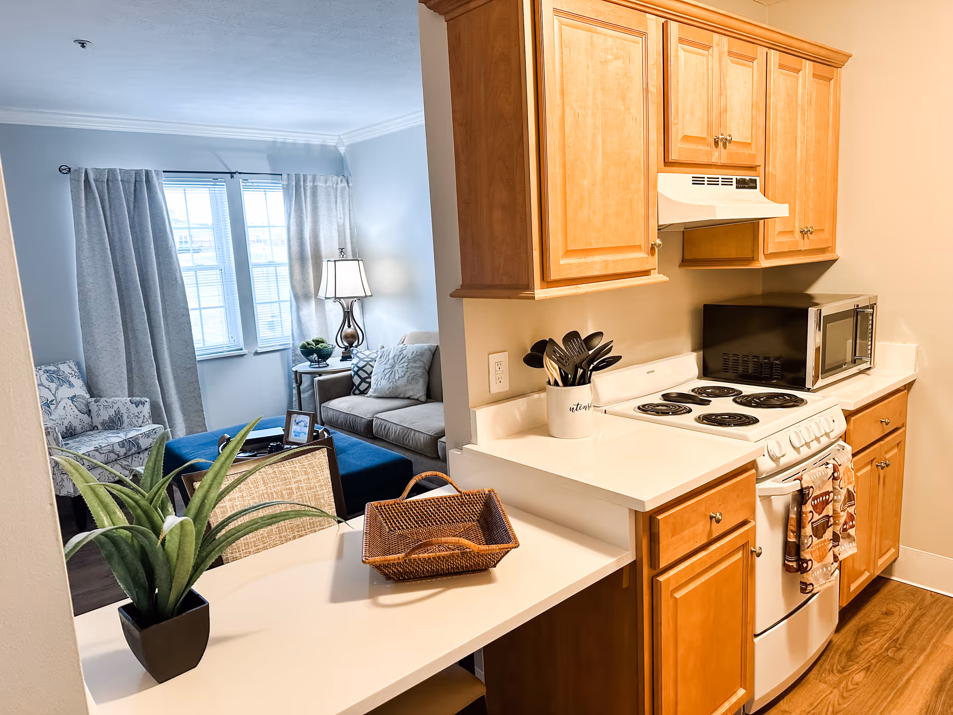 Small kitchen with wooden cabinets, white countertops and a stove opening to a cozy living room with a sofa and lamp.