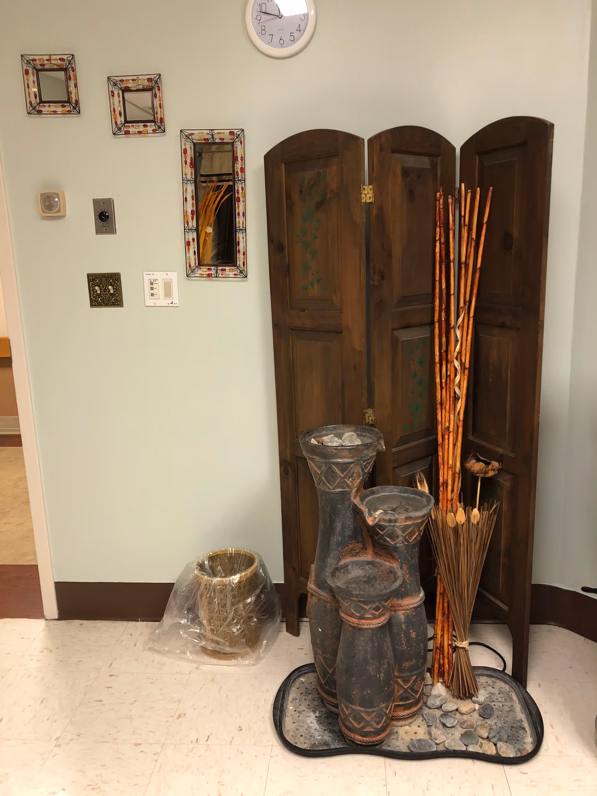 A corner of an interior room with a decorative wooden folding screen, tall vases with dried sticks and reeds, a small pot wrapped in plastic on the floor, three small decorative mirrors on the wall, a clock, and various wall switches and controls.