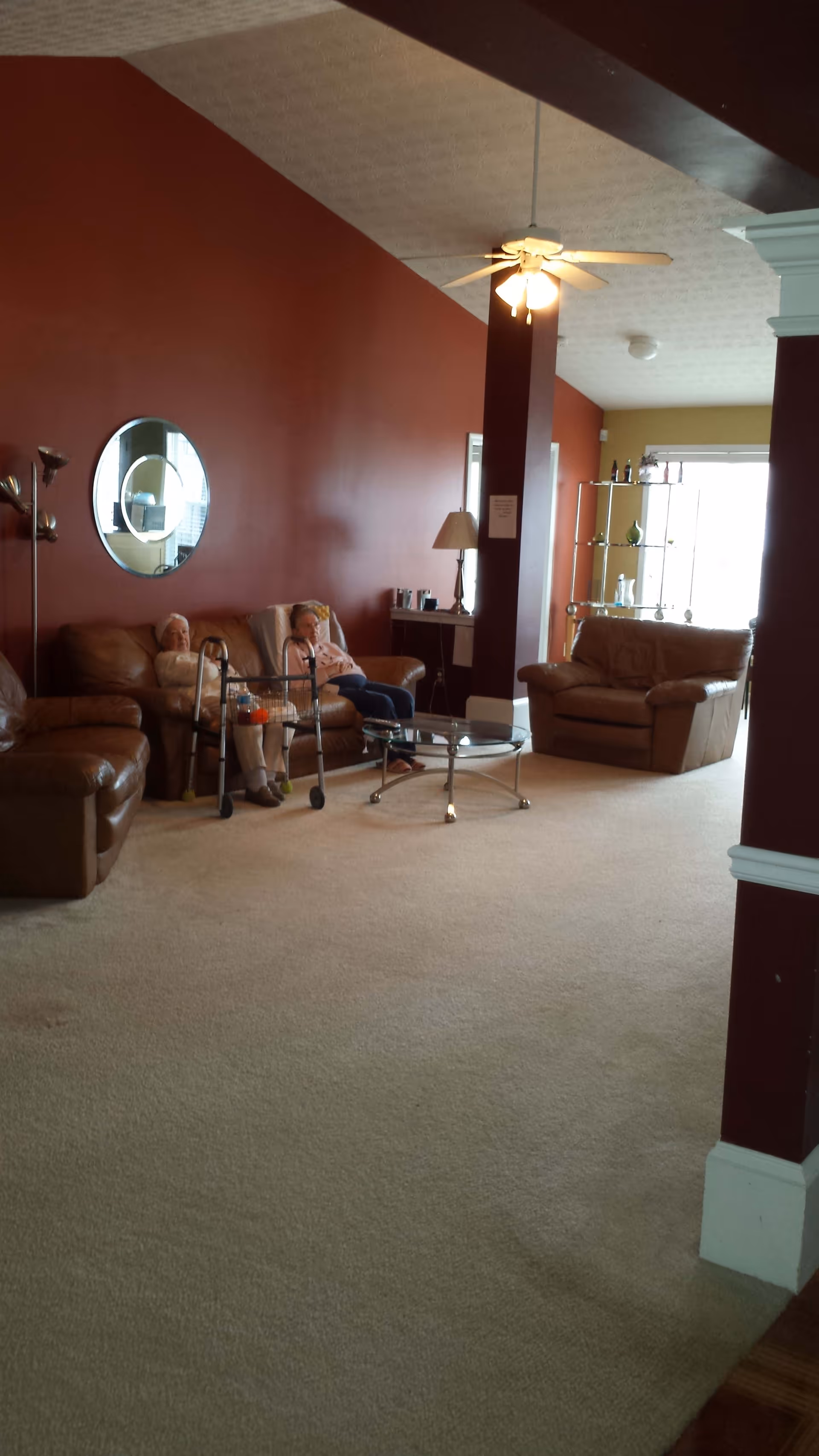 A living room with beige carpet, brown leather sofas, a glass coffee table, and a ceiling fan. Two elderly women are seated on one of the sofas, with a walker in front of them. The walls are painted in a deep red color, and there is a round mirror on the wall behind the women. A lamp and a shelving unit with decorative items are visible in the background.