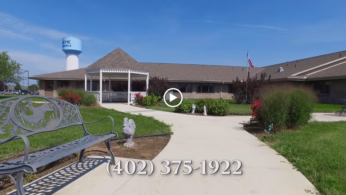 Exterior view of a single-story care facility with a gazebo, paved walkway, bench, and lawn with a phone number overlay.