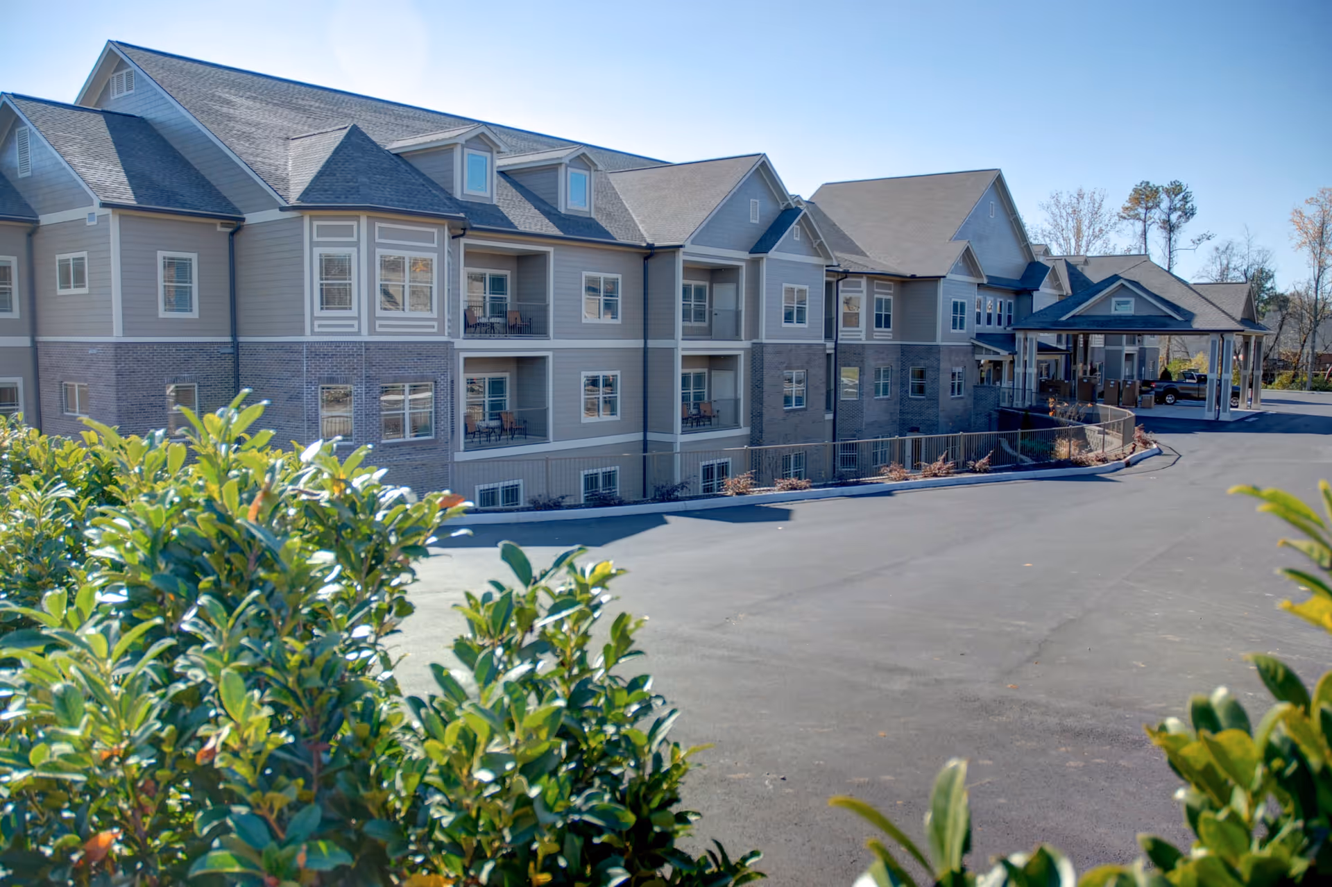Exterior view of a multi-story senior living facility building with balconies, a covered entrance, and a paved driveway, partially framed by green bushes in the foreground under a clear blue sky.
