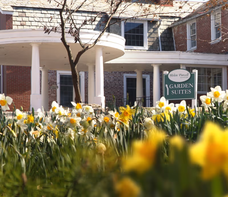 Front entrance of a brick senior living building with white columns, a 'Garden Suites' sign and daffodils in the foreground.