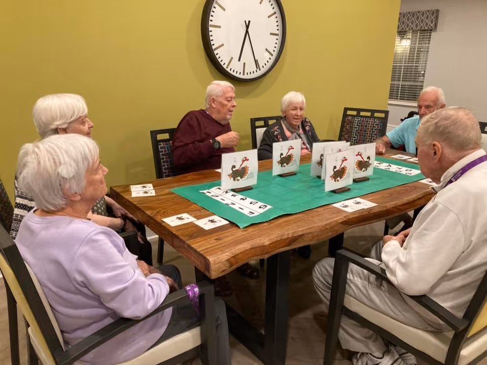 A group of six elderly people sitting around a wooden table playing a card game with green felt and card holders, in a room with yellow walls and a large clock on the wall.