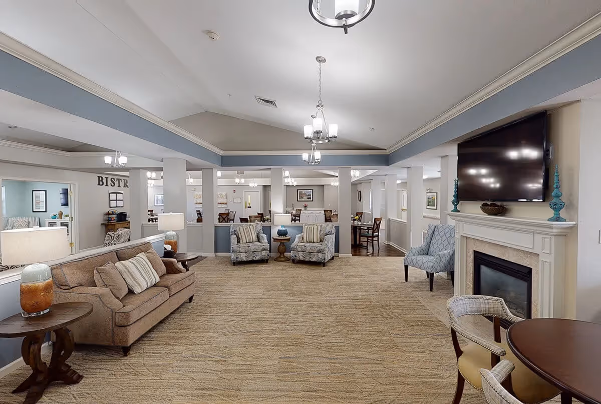 A spacious and well-lit living room area in Chandler Place featuring a beige sofa with striped pillows, two patterned armchairs with a small round table between them, a fireplace with a mounted flat-screen TV above it, and additional seating with a round table and chairs. The room has a beige carpet, light-colored walls with blue trim, and multiple ceiling light fixtures. In the background, there is a dining area visible with tables and chairs.