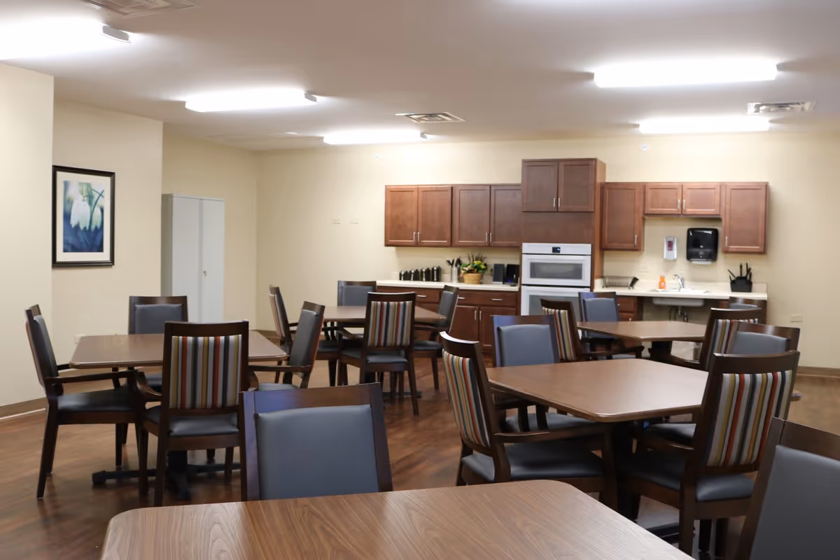 Community dining area with several wooden tables and upholstered chairs and a small kitchen with cabinets and appliances along the back wall.