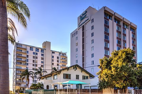 Tall white senior living building with a small front house, palm trees, and turquoise umbrellas.