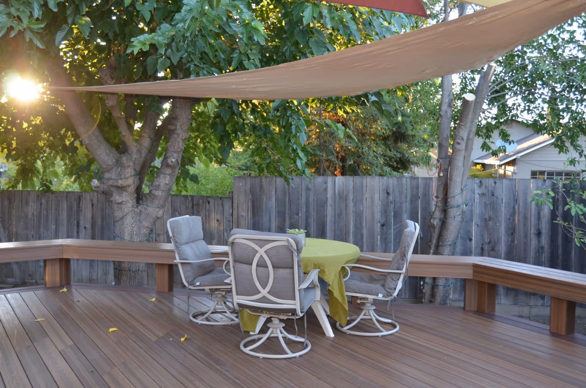 Outdoor wooden deck area with a round table covered by a green tablecloth and surrounded by four cushioned swivel chairs. There is a wooden bench along the perimeter of the deck, trees providing shade, and a wooden fence in the background. Sunlight filters through the leaves.