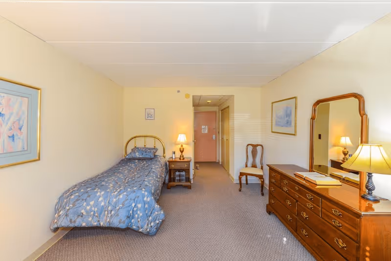 A simple bedroom with a single bed covered in a blue patterned comforter, a wooden nightstand with a lamp, a wooden dresser with a mirror and a lamp, a wooden chair, and framed artwork on the walls. The room has beige walls and carpeted floor, with a door visible at the far end.