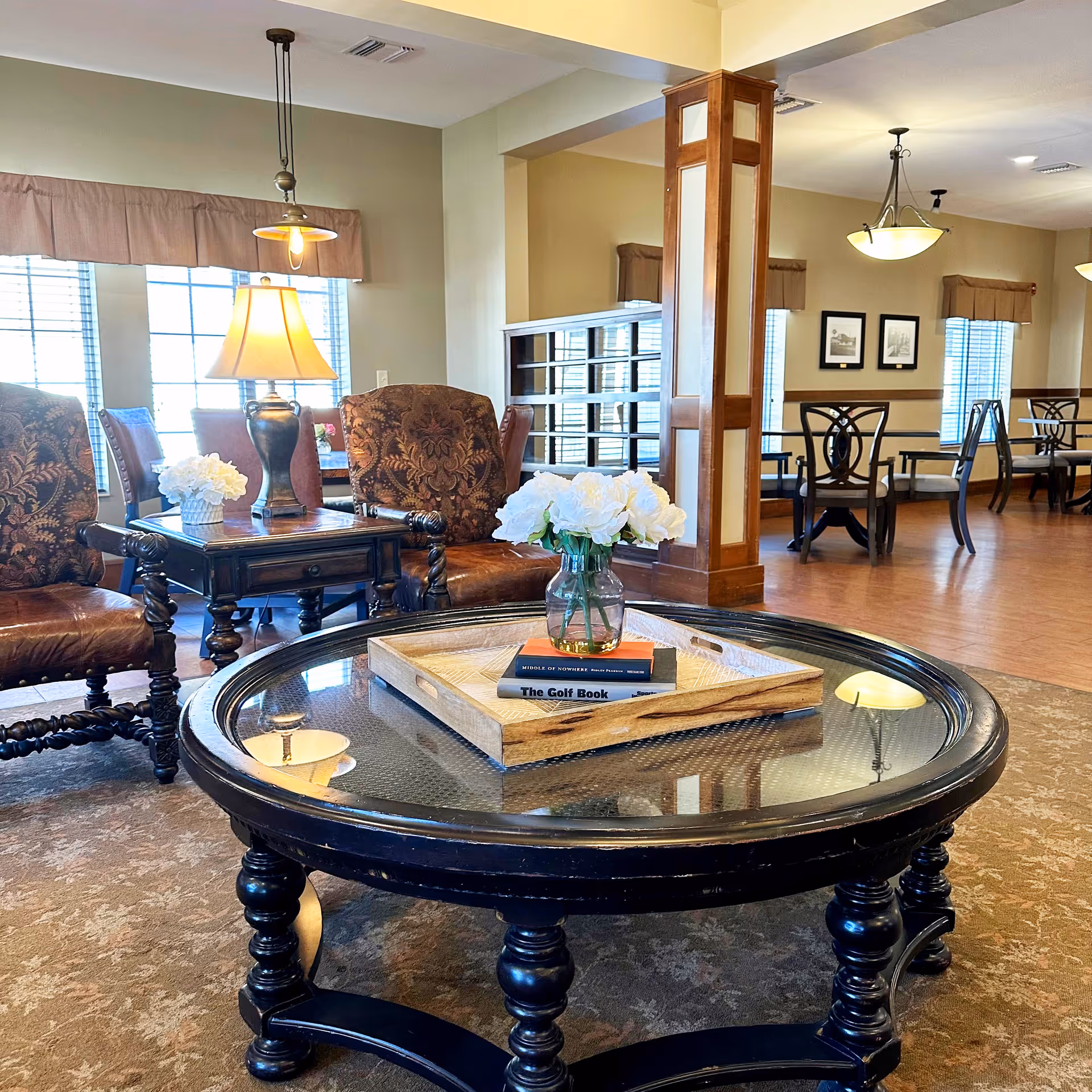 A cozy senior living common area with a round glass-top coffee table holding a wooden tray with white flowers and books. Surrounding the table are upholstered chairs with ornate wooden legs. In the background, there are more chairs and tables near large windows with blinds and valances, and warm lighting fixtures hanging from the ceiling.