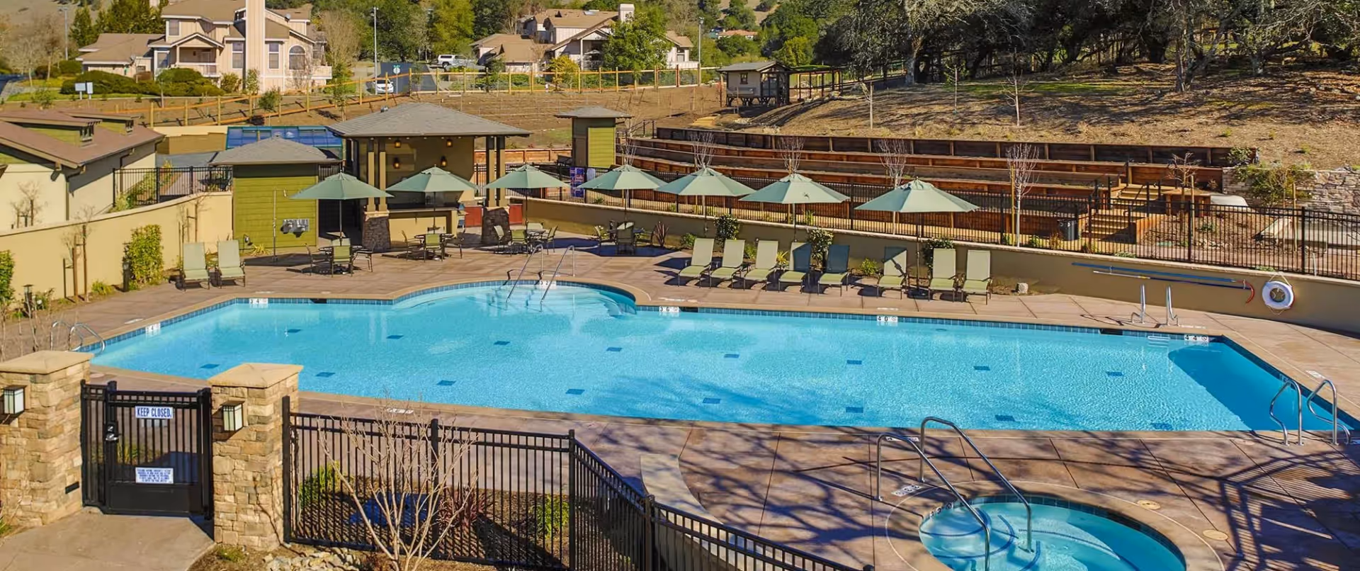 Outdoor swimming pool area with lounge chairs and umbrellas arranged around the pool. There is a smaller circular hot tub in the foreground and a gated entrance with stone pillars. Trees and residential buildings are visible in the background.