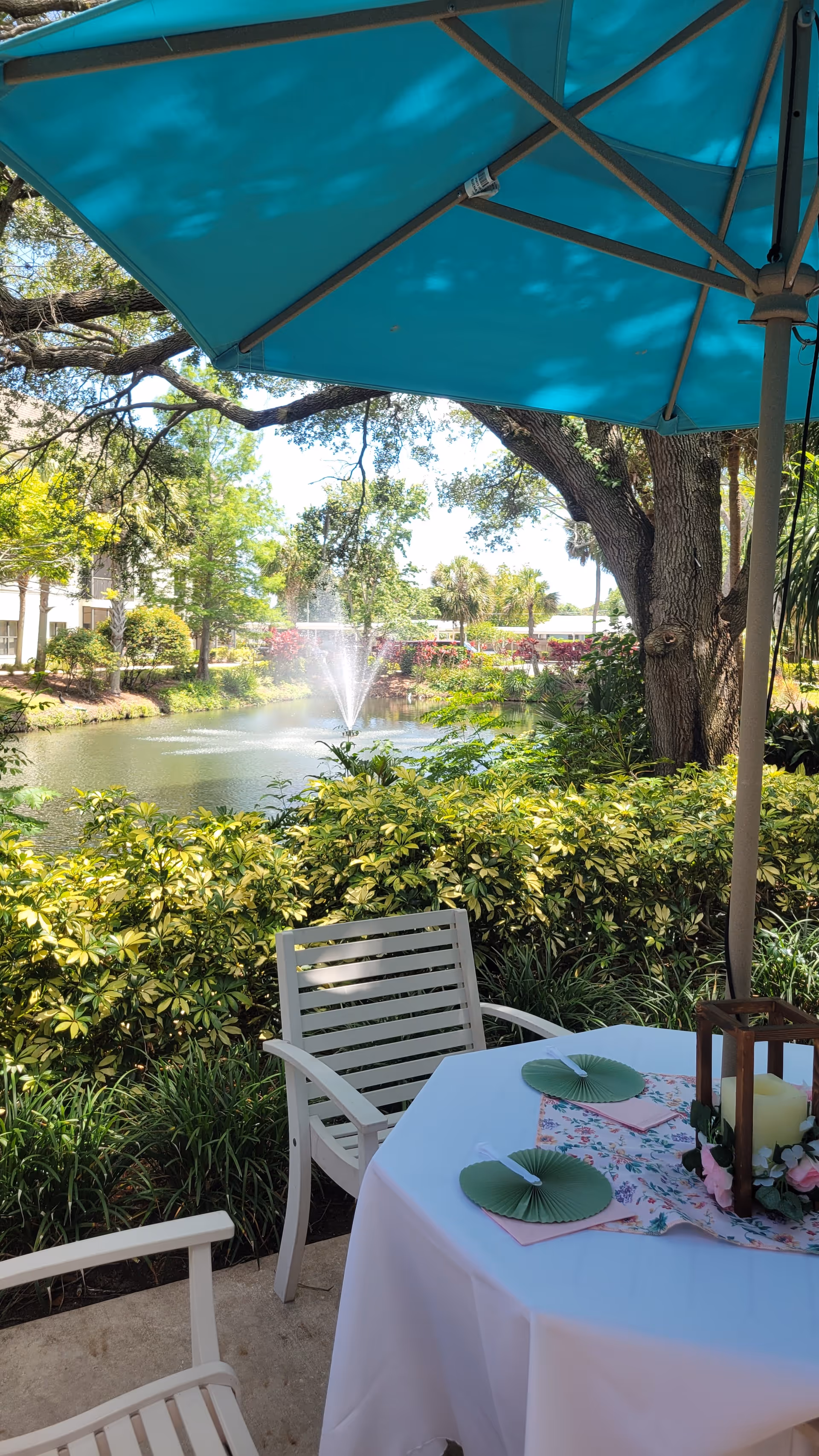 Outdoor seating area with a white table covered with a white tablecloth and floral centerpiece, two white chairs, and a large blue umbrella. In the background, there is a pond with a water fountain, surrounded by green bushes and trees under a sunny sky.