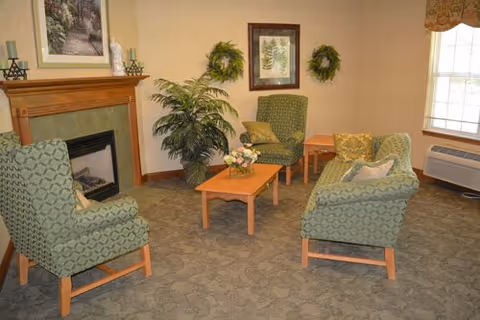 A cozy living room area with four green patterned armchairs arranged around two wooden coffee tables. The room features a fireplace with decorative items on the mantel, two wreaths and a framed botanical print on the wall, a large potted plant, and a window with a valance letting in natural light.