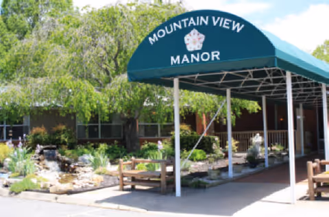 Entrance of Mountain View Manor Nursing Center with a green canopy displaying the facility name. The area features a landscaped garden with rocks, plants, and benches under a sunny sky.