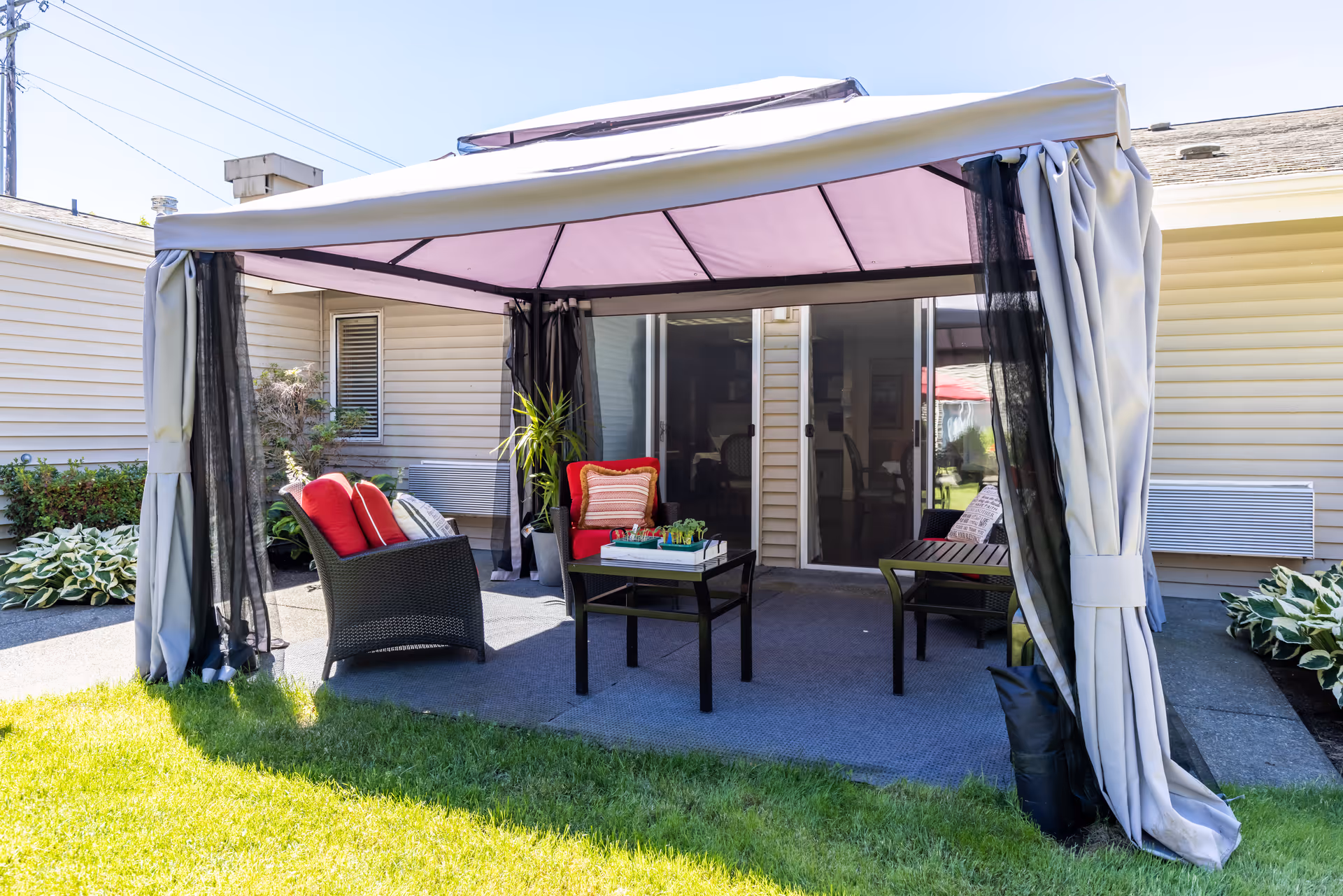 A shaded backyard patio with a gazebo, wicker seating with red cushions, a coffee table, and sliding glass doors leading into the building.
