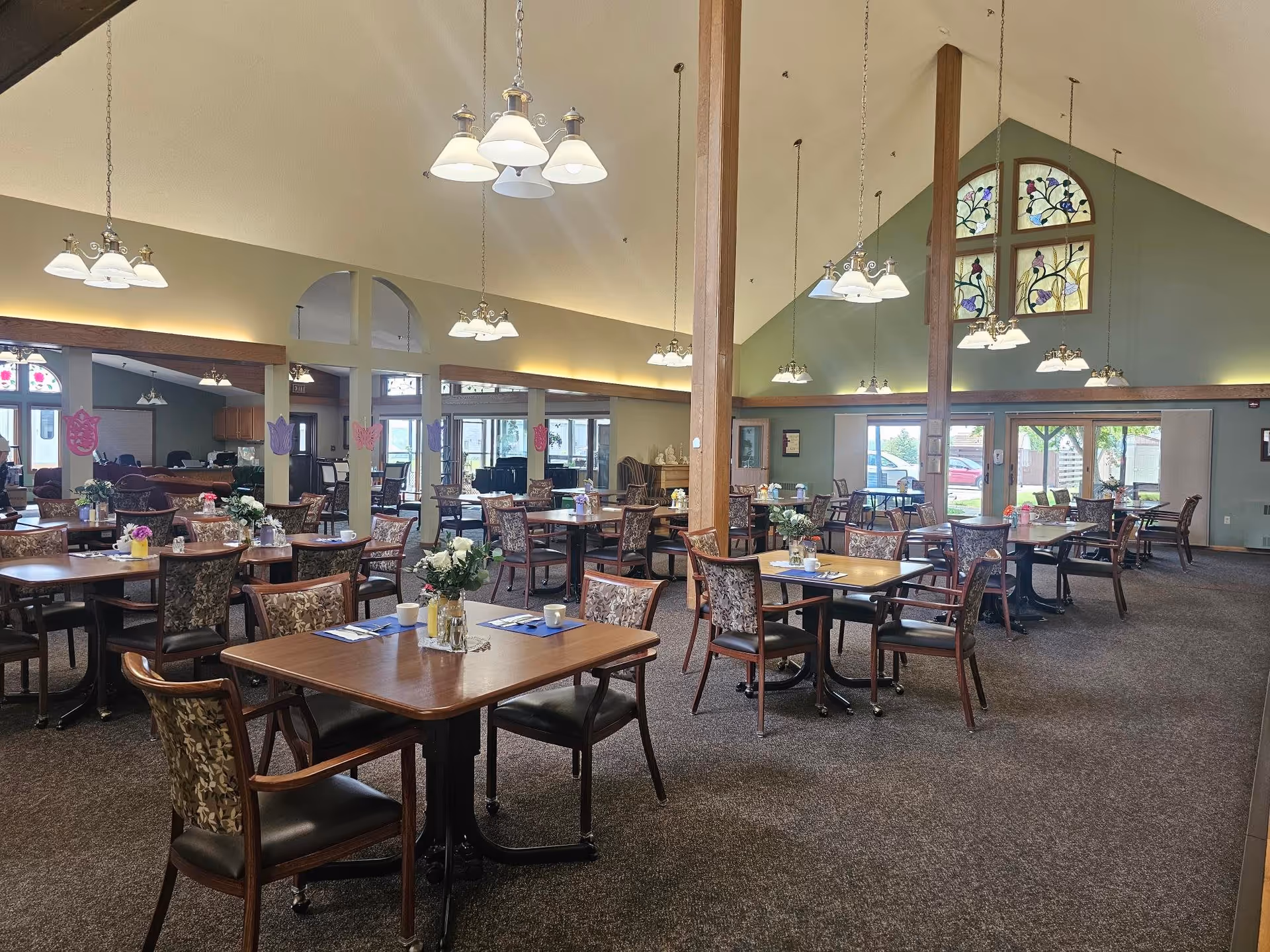 Spacious dining room in an assisted living facility with multiple wooden tables and chairs arranged neatly. Each table has a small floral centerpiece and place settings. The room features high vaulted ceilings with hanging light fixtures and large windows allowing natural light to enter. Decorative stained glass windows are visible on one wall.