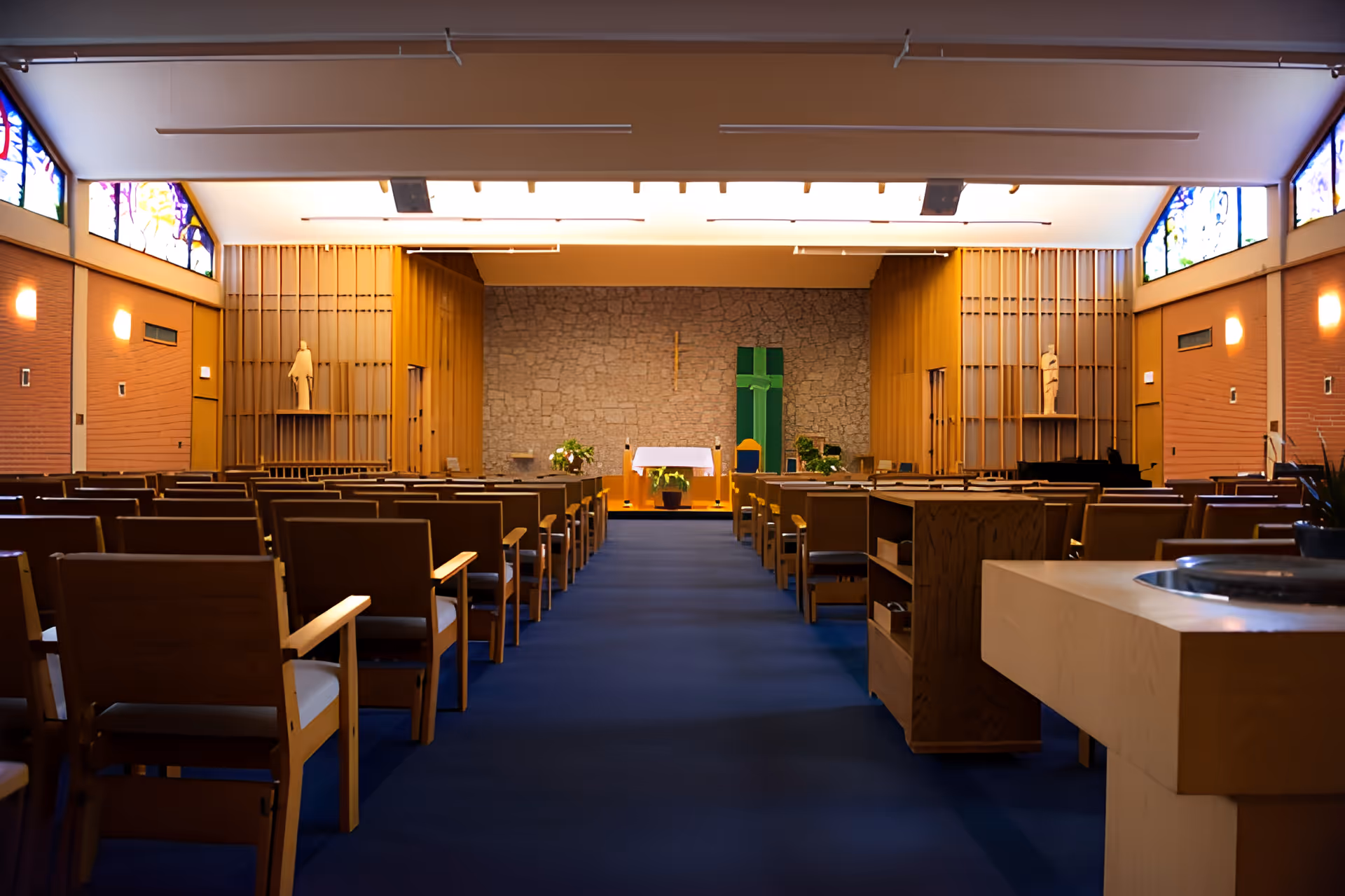 Interior view of a chapel or worship room with rows of wooden chairs facing an altar at the front. The altar is set against a stone wall with a cross and green cloth decoration. The room has stained glass windows near the ceiling and warm lighting on the walls.