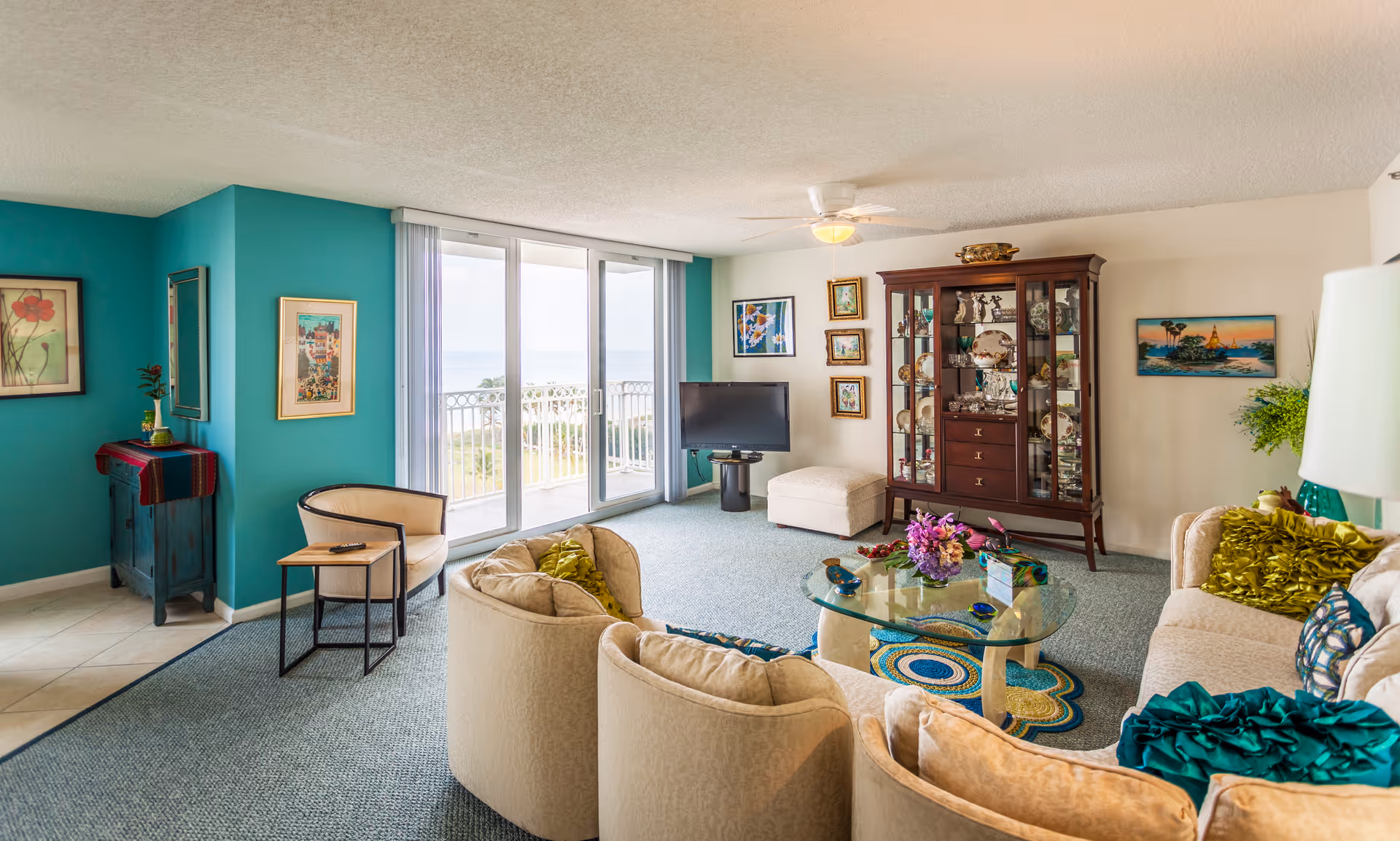 A bright and spacious living room with a curved beige sofa adorned with green and blue cushions, a glass coffee table with decorative items, a wooden display cabinet filled with china, and a flat-screen TV near sliding glass doors that open to a balcony with a view of greenery and water. The walls are painted white and teal, decorated with framed artwork, and the floor is carpeted.