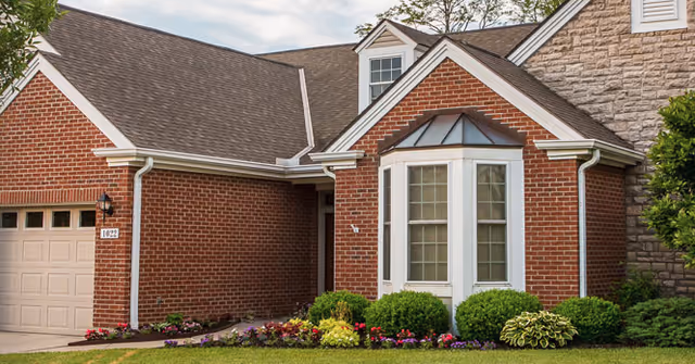 Exterior view of a brick building with a garage door, a bay window, and a small garden with various shrubs and flowers in front.