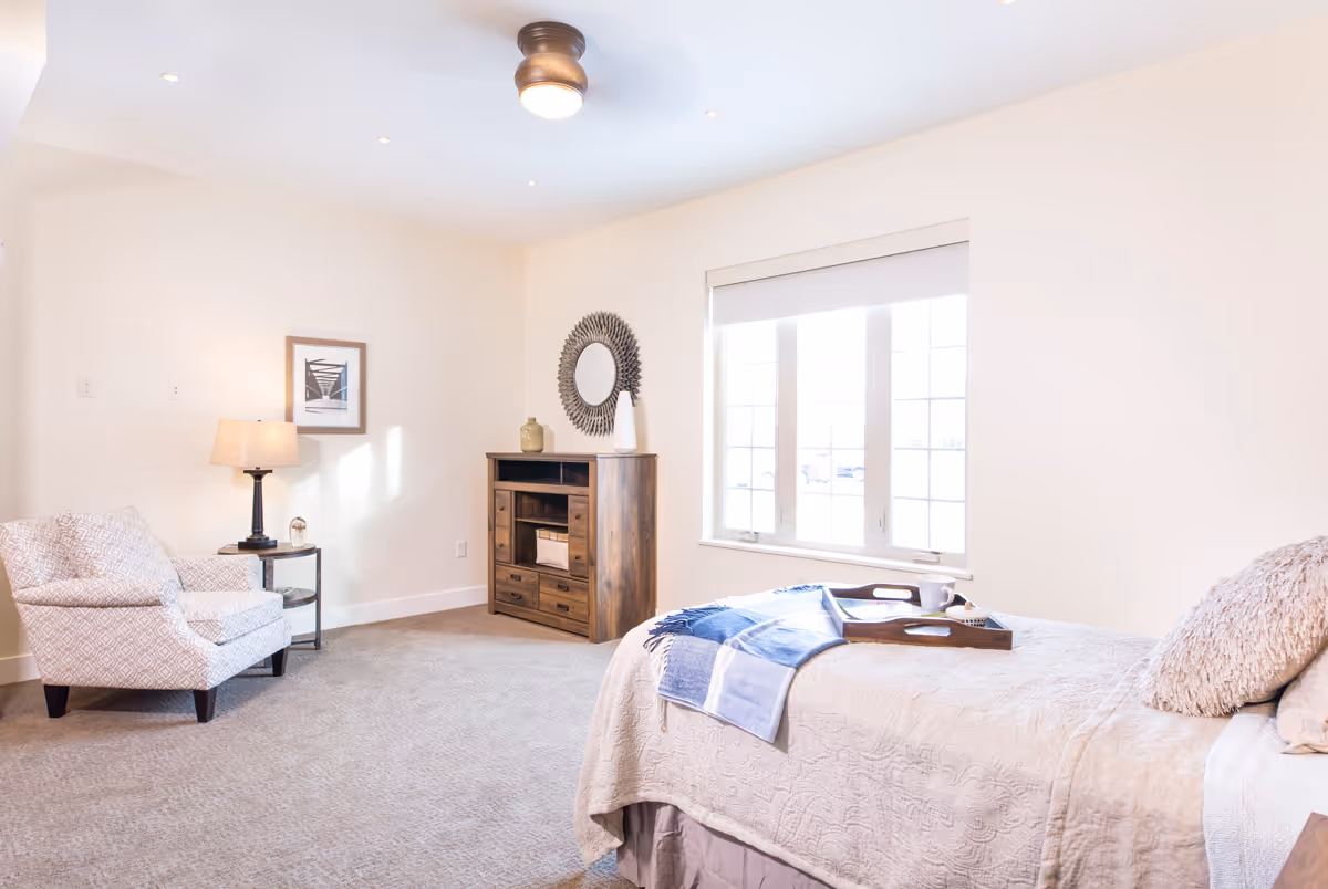 A bright and cozy bedroom with a large window letting in natural light. The room features a bed with a beige quilt, a blue throw blanket, and a tray with a cup on it. There is a patterned armchair next to a small round side table with a lamp. A wooden dresser with drawers and a decorative round mirror above it is positioned against the wall.