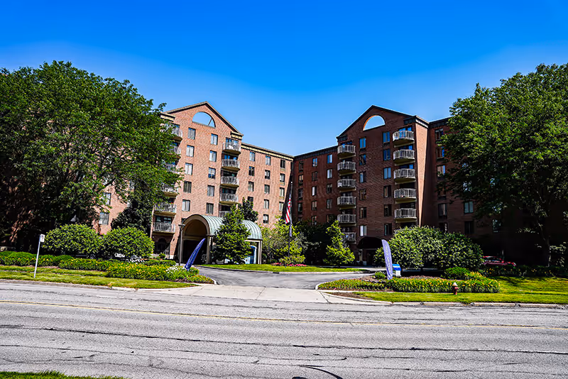 Front exterior of a multi-story brick senior living building with a covered entrance, balconies, and landscaped grounds.