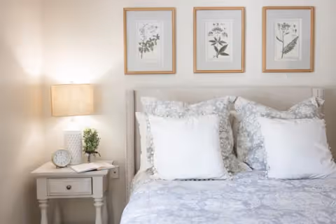 A neatly made bed with white and patterned pillows and a matching bedspread. Above the bed are three framed botanical prints. To the left of the bed is a white nightstand with a table lamp, a small plant, a clock, and an open book.