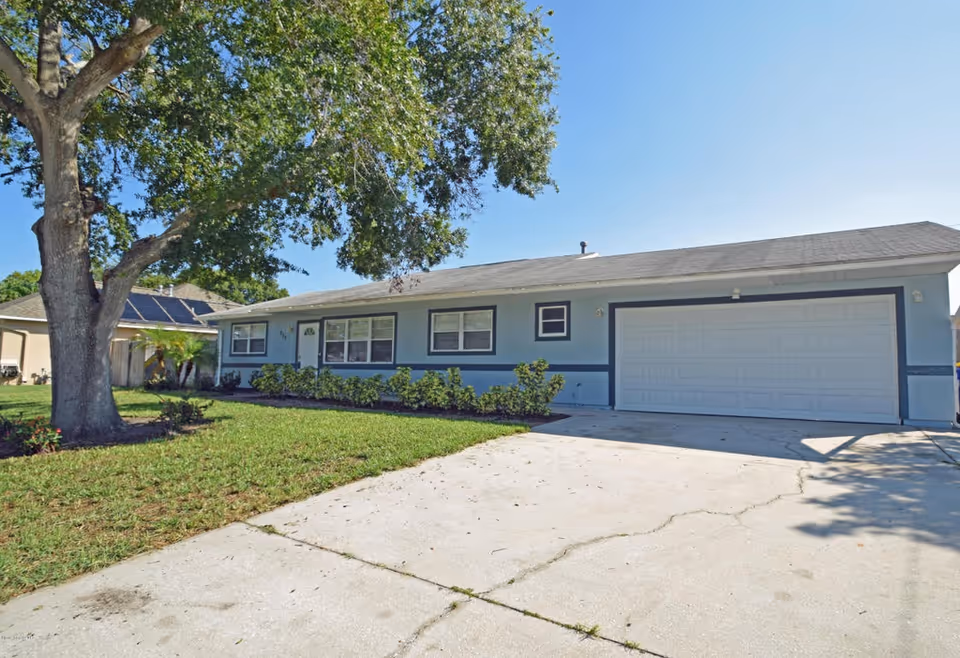 Single-story light-blue house with an attached two-car garage, front lawn, and a large tree by the driveway.