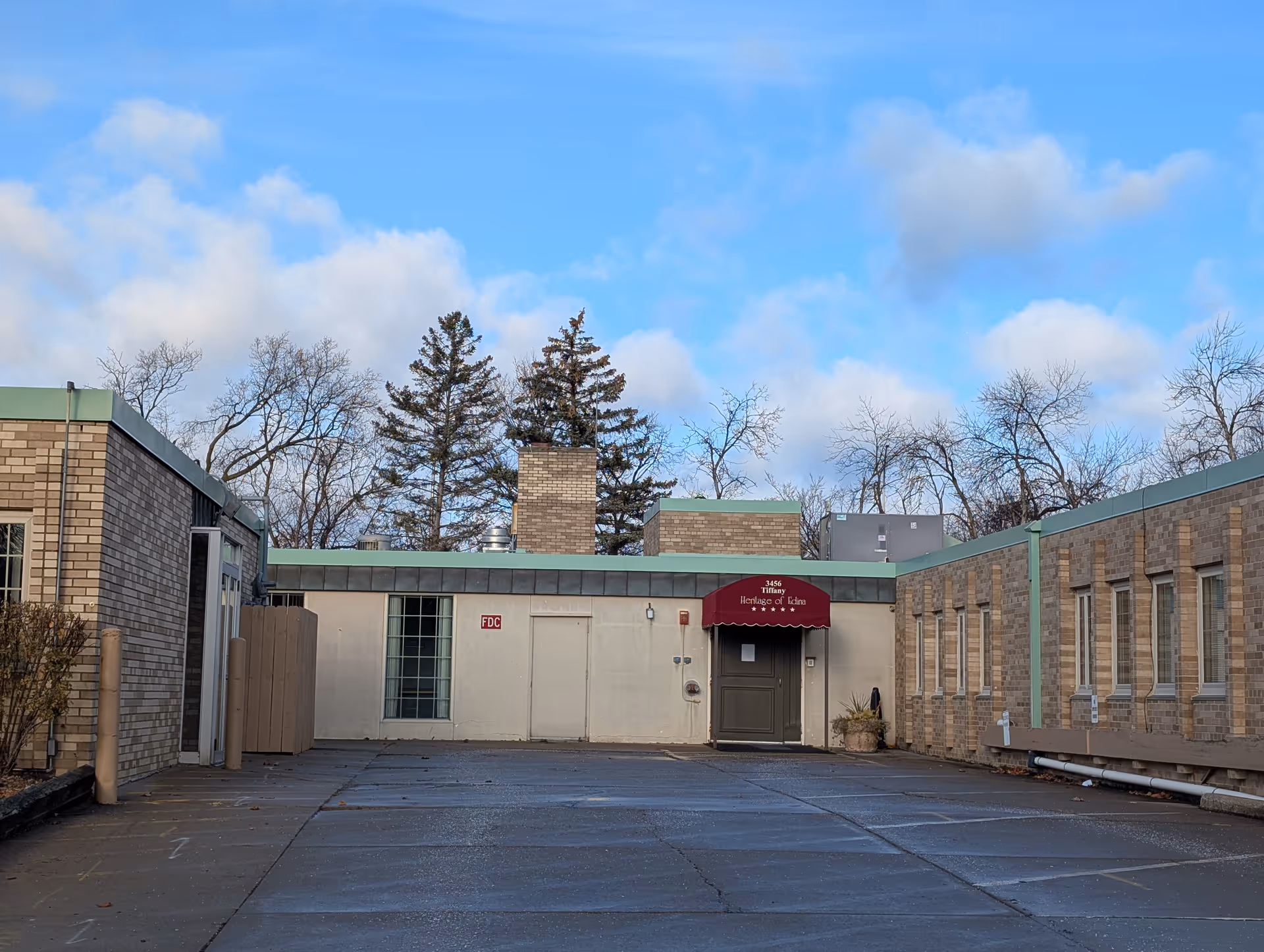 Exterior view of a single-story brick building with a flat roof and a maroon awning above a door that reads '3456 Tiffany Heritage of Edina'. The building is surrounded by leafless trees under a partly cloudy blue sky.