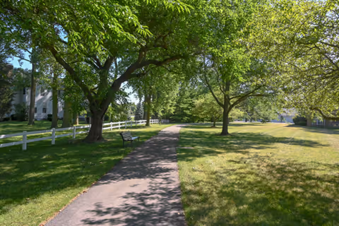 Tree-lined paved walkway with benches and a white fence running through a grassy lawn on a sunny day at The Parkvue Community.