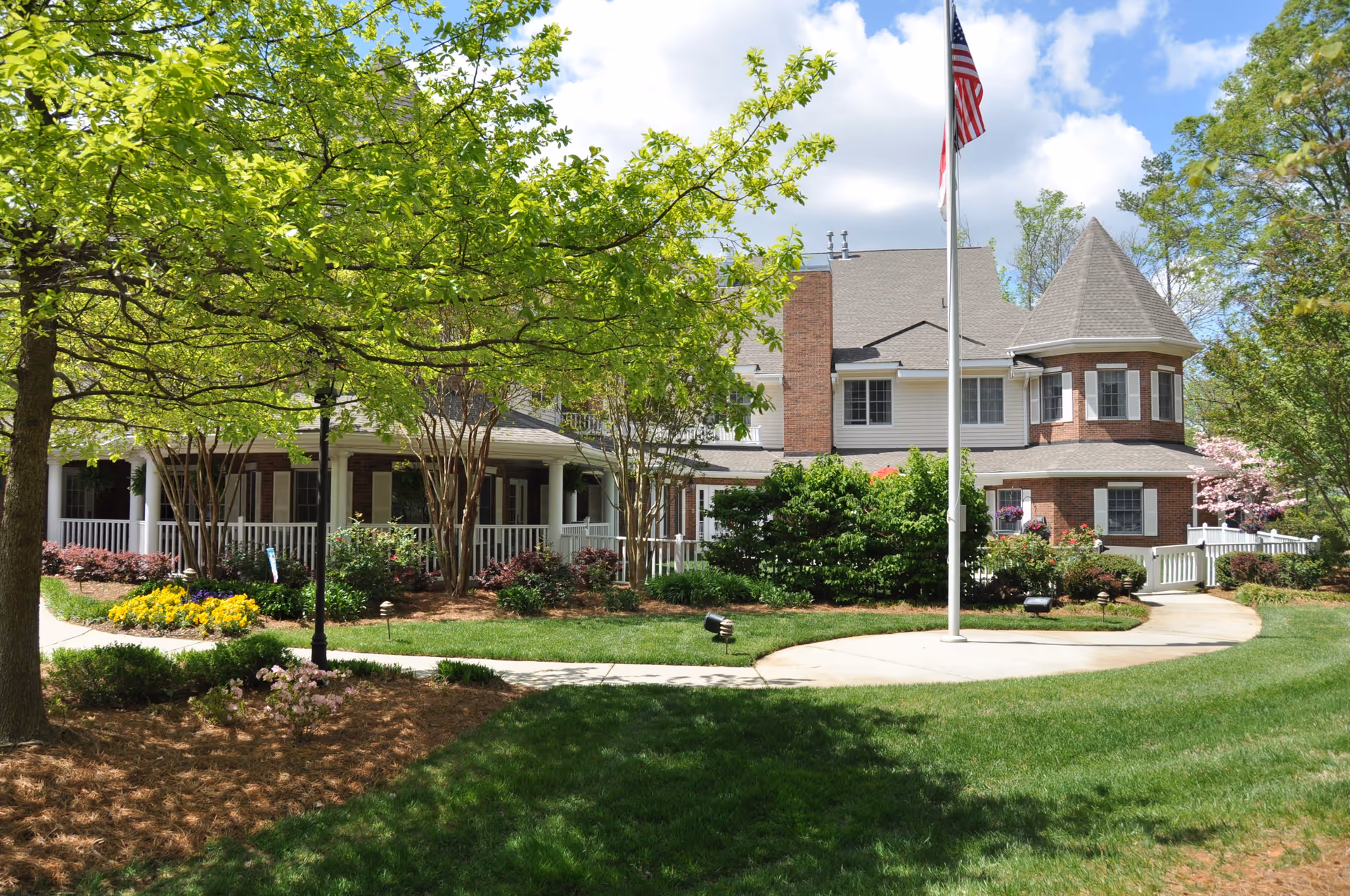 Exterior view of a senior living facility building with a well-maintained lawn, trees, shrubs, and flower beds. The building has a porch with white railings, brick and siding exterior, and a turret-like architectural feature. An American flag is flying on a flagpole in front of the building under a partly cloudy sky.
