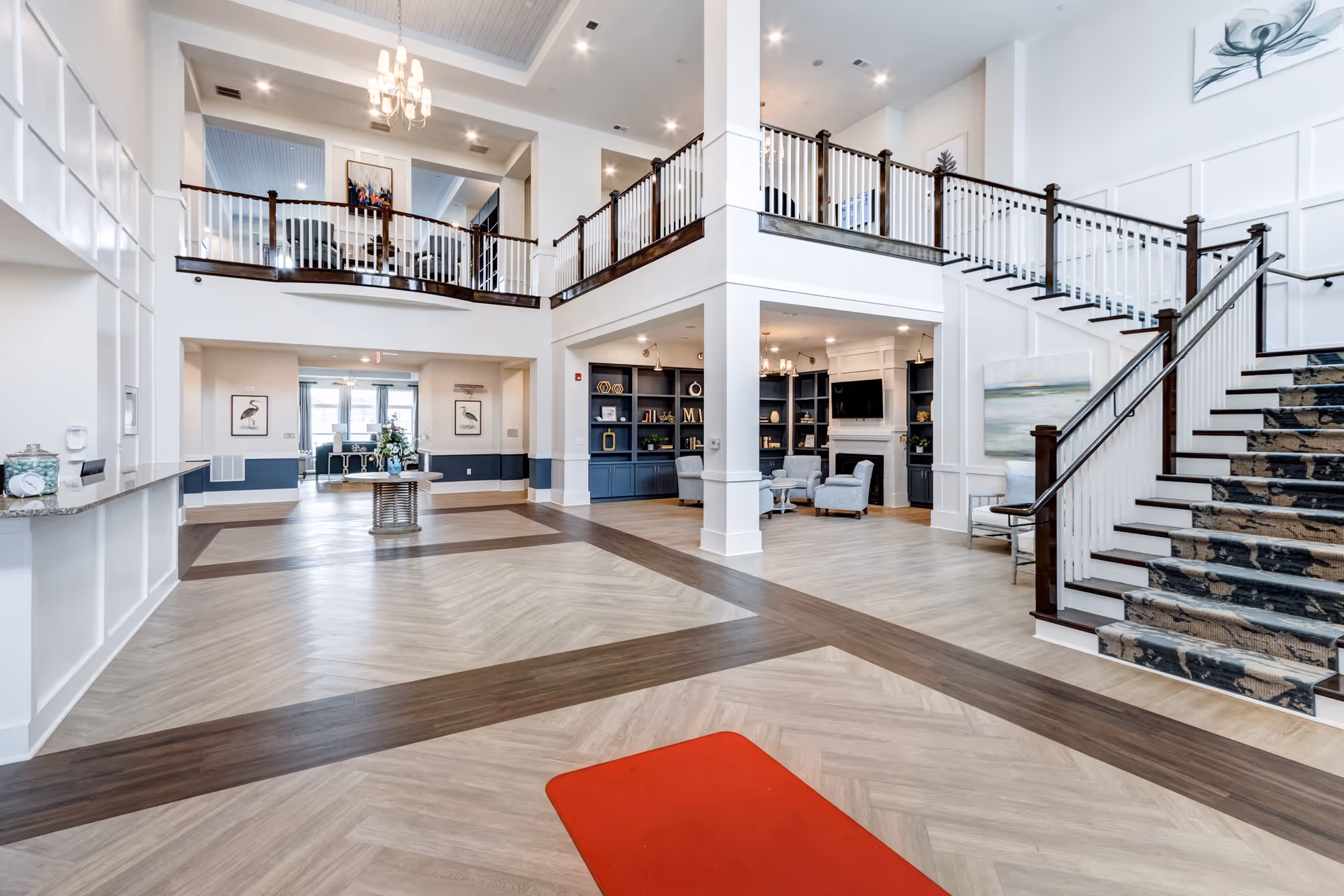 Spacious and bright senior living facility lobby with high ceilings, a chandelier, a staircase with patterned carpet, seating area with chairs and a fireplace, decorative shelving, and a table with flowers in the center.
