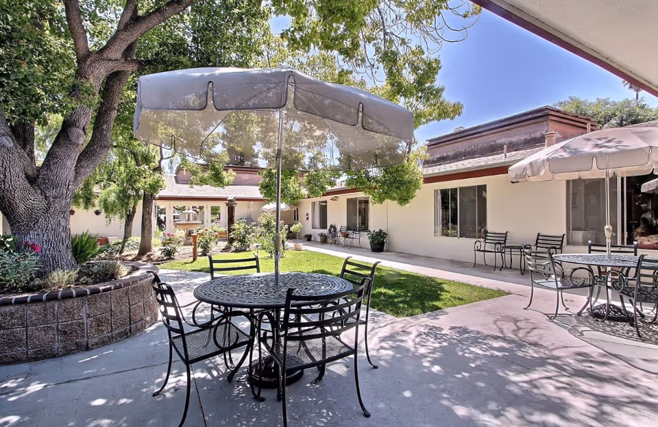 Outdoor courtyard area at The Vineyards Healthcare Center featuring metal tables and chairs with umbrellas, surrounded by trees, plants, and a building with large windows.