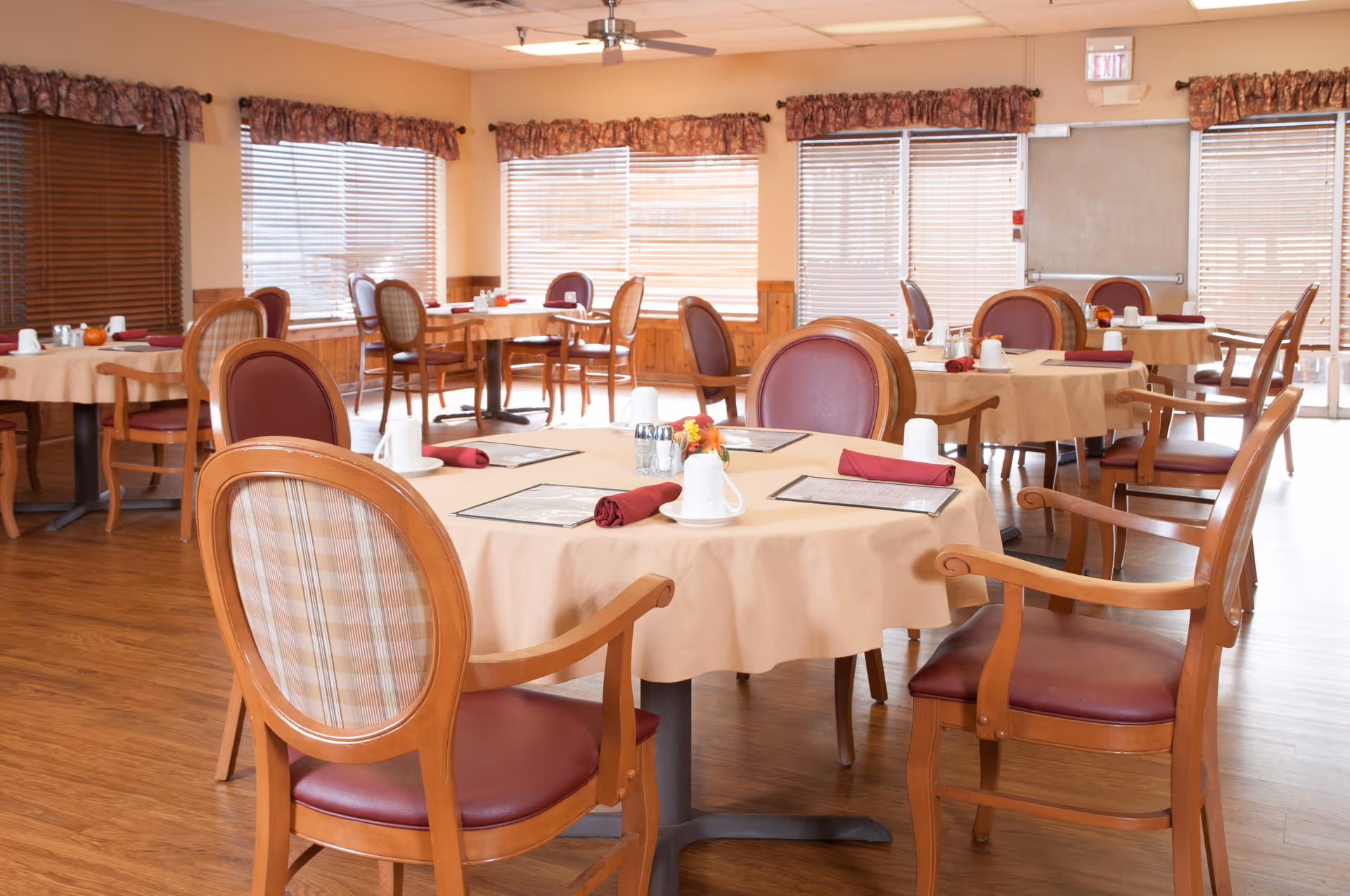 Bright dining room with round tables covered in beige tablecloths, place settings, and wooden chairs.