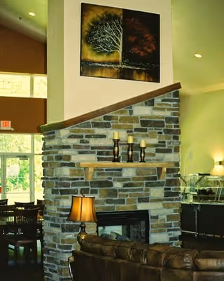 Interior view of a living room area featuring a stone fireplace with a wooden mantel holding three candles. Above the fireplace hangs a painting of a tree with one half in green and the other half in red. In front of the fireplace is a brown leather sofa and a table lamp. In the background, there are dining tables and chairs near large windows letting in natural light.