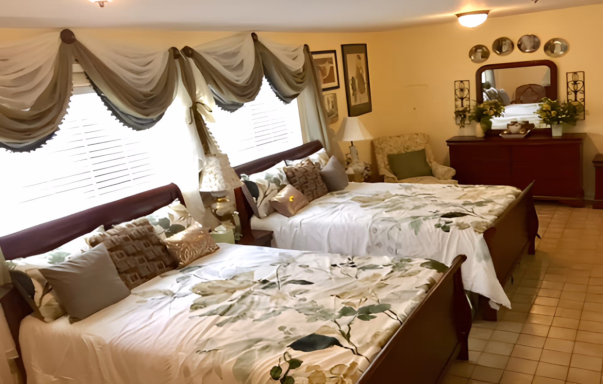 A cozy bedroom with two beds featuring floral bedspreads and multiple pillows. The room has a large window with decorative drapes, a cushioned armchair with a green pillow, a wooden dresser with a mirror, and various framed artworks on the walls. The floor is tiled and the lighting is warm.