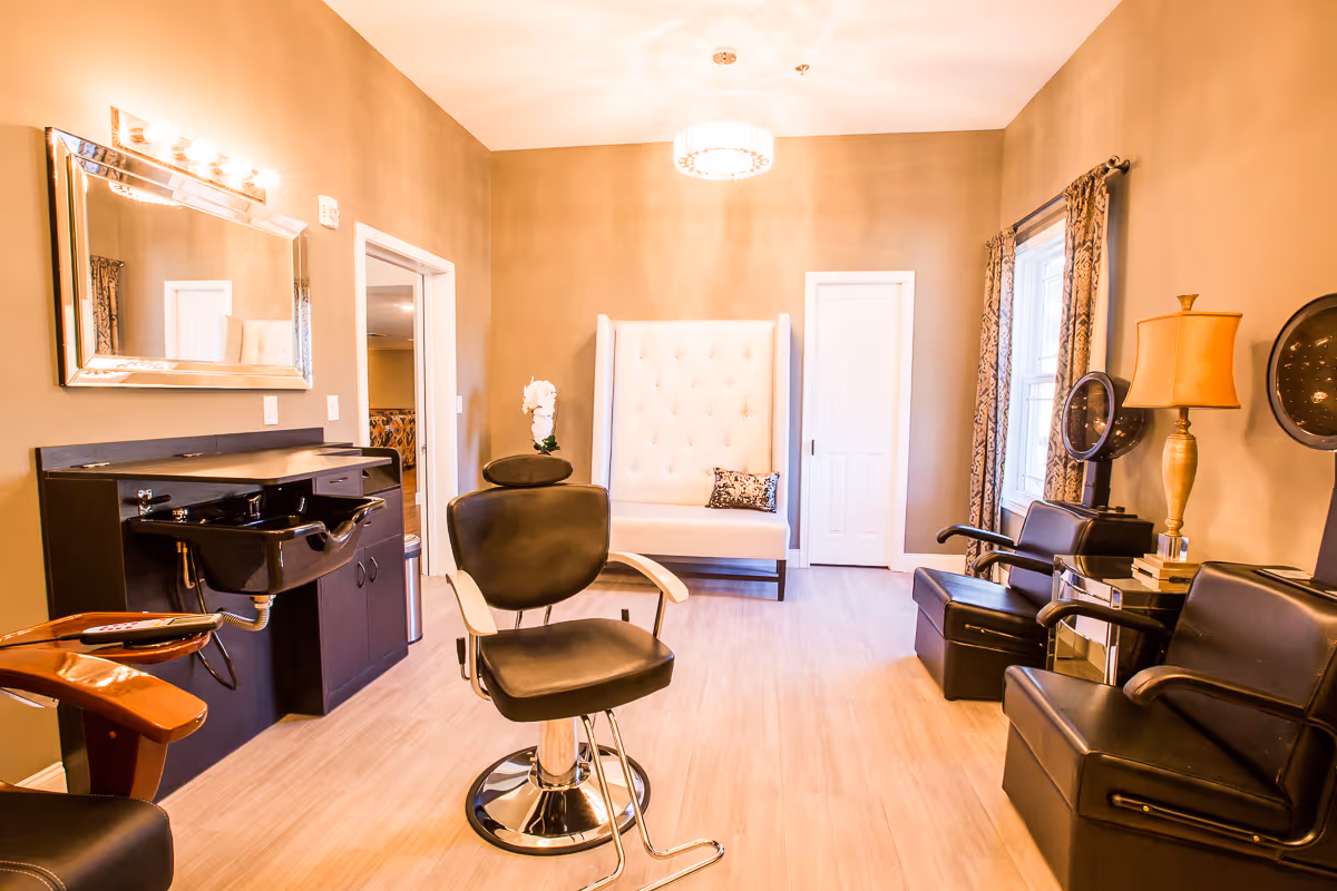 Interior of a salon room with a black salon chair in the center, a black hair washing station with a mirror on the left wall, two black salon chairs with hair dryers on the right side, a beige tufted bench with a decorative pillow against the back wall, and a window with patterned curtains. The room has beige walls and light wood flooring.
