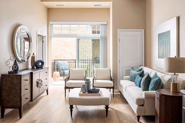 Bright living room with a beige sofa adorned with teal and blue pillows, two matching armchairs, and a cushioned ottoman in the center. A wooden sideboard with decorative items and a round mirror hangs on the left wall. Sliding glass doors lead to a small balcony with outdoor seating. Light wood flooring and neutral-colored walls complete the space.