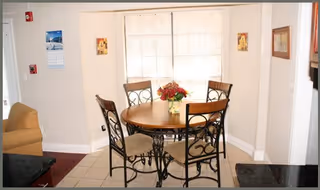 A small dining area with a round wooden table and four metal chairs with cushioned seats. A vase with red flowers is placed in the center of the table. The dining area is next to a large window with blinds, and part of a beige armchair and kitchen counter are visible.