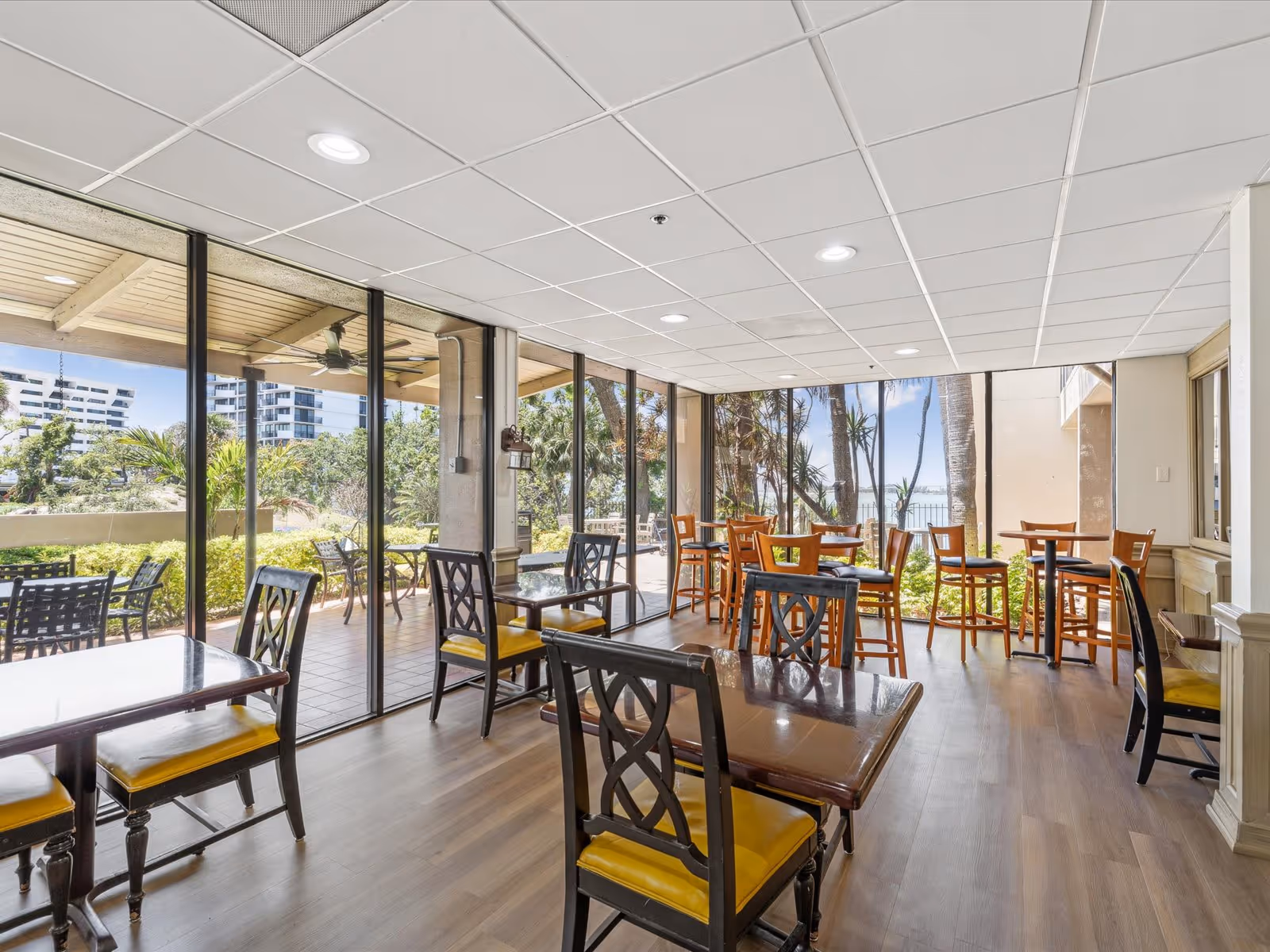 A bright dining area with multiple tables and chairs featuring yellow cushions. Large floor-to-ceiling windows provide a view of an outdoor patio with additional seating and greenery. The ceiling has recessed lighting, and the floor is wooden.