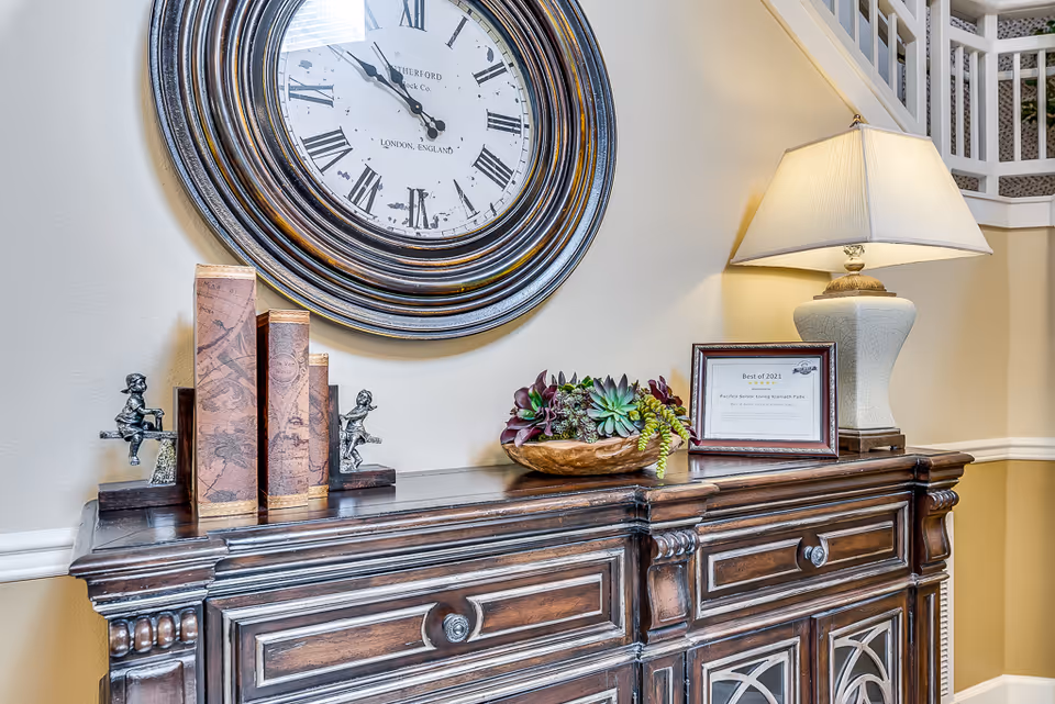 A decorative wooden console table topped with a lamp, framed award, books, and a succulent arrangement beneath a large round wall clock.