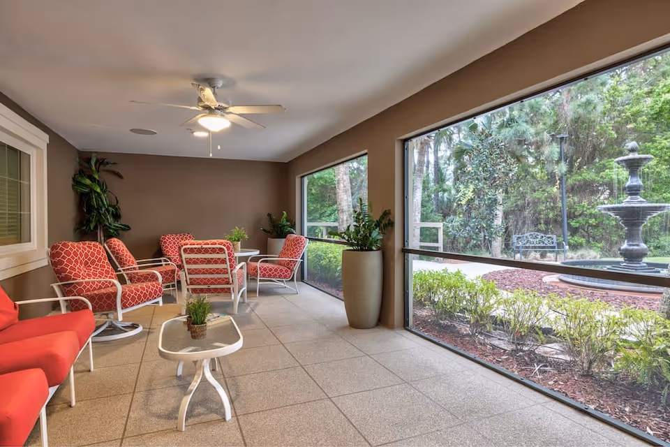 A covered patio area with red cushioned chairs and a small glass-top table, overlooking a garden with a black tiered fountain and greenery. The patio has large screened windows and potted plants, with a ceiling fan above.