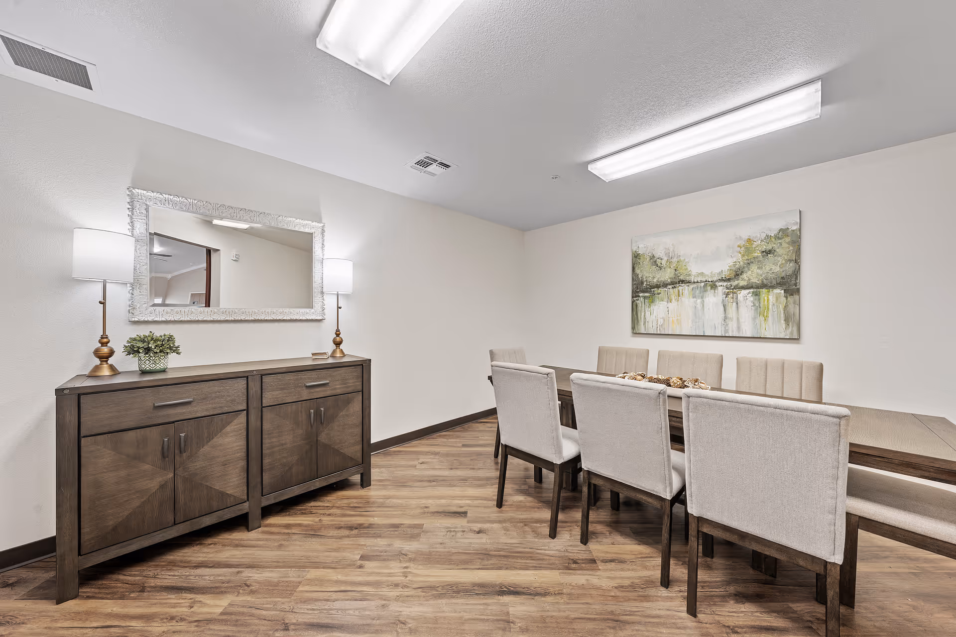 A dining room with a wooden dining table surrounded by six beige upholstered chairs. On the left side, there is a wooden sideboard with two lamps and a small plant on top. A large mirror hangs above the sideboard, and a landscape painting is mounted on the wall above the dining table. The room has wood flooring and white walls with bright overhead lighting.