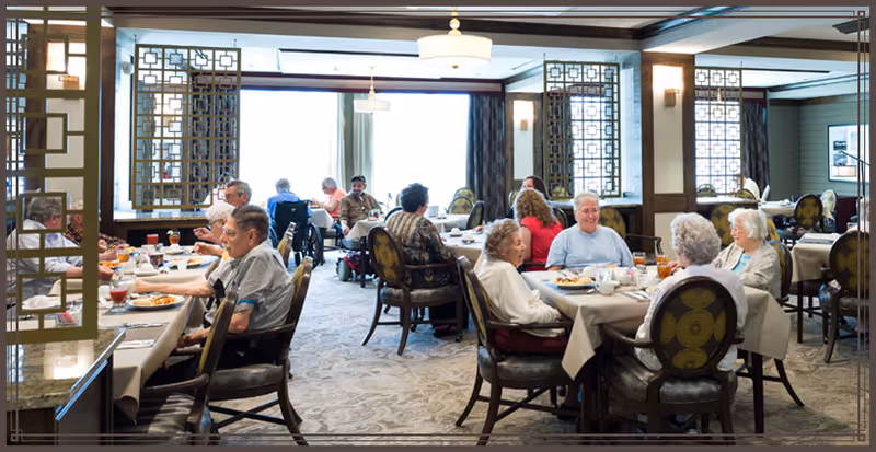 A group of elderly people seated at multiple tables in a well-lit dining room, enjoying meals and socializing. The room features decorative wooden partitions, carpeted floors, and large windows with curtains allowing natural light to enter.