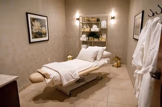 A serene spa treatment room with a padded massage table covered in white towels and pillows. The room has soft lighting from wall sconces, a shelf stocked with rolled towels and spa products, framed floral artwork on the wall, and two white robes hanging on hooks by the door.