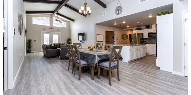 Open-plan interior of a senior living common area with a dining table, kitchen, and seating area under vaulted ceilings.