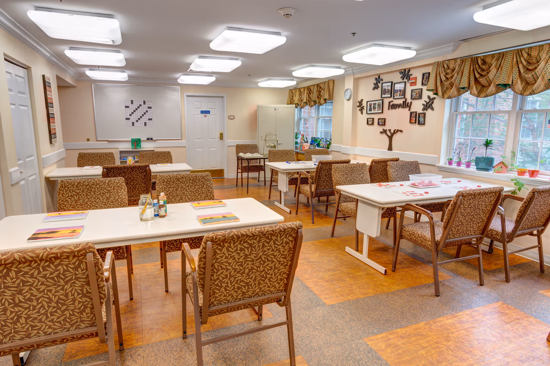 A well-lit activity room with several tables and chairs arranged for group activities. The tables have various materials on them, including papers and small containers. The walls are decorated with family-themed pictures and a clock. Large windows with patterned valances let in natural light, and the floor has a mix of carpet and wood-like tiles.
