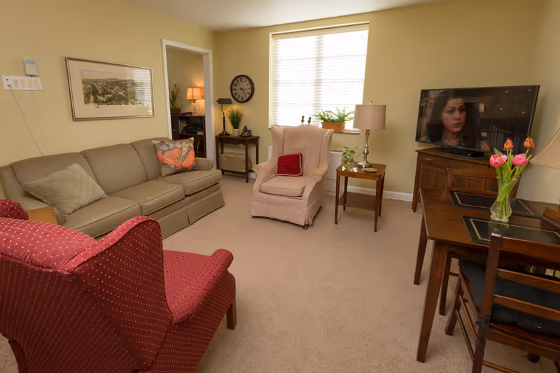 A cozy living room with a sofa and two armchairs, a TV on a cabinet, a small dining table, and a window with blinds.