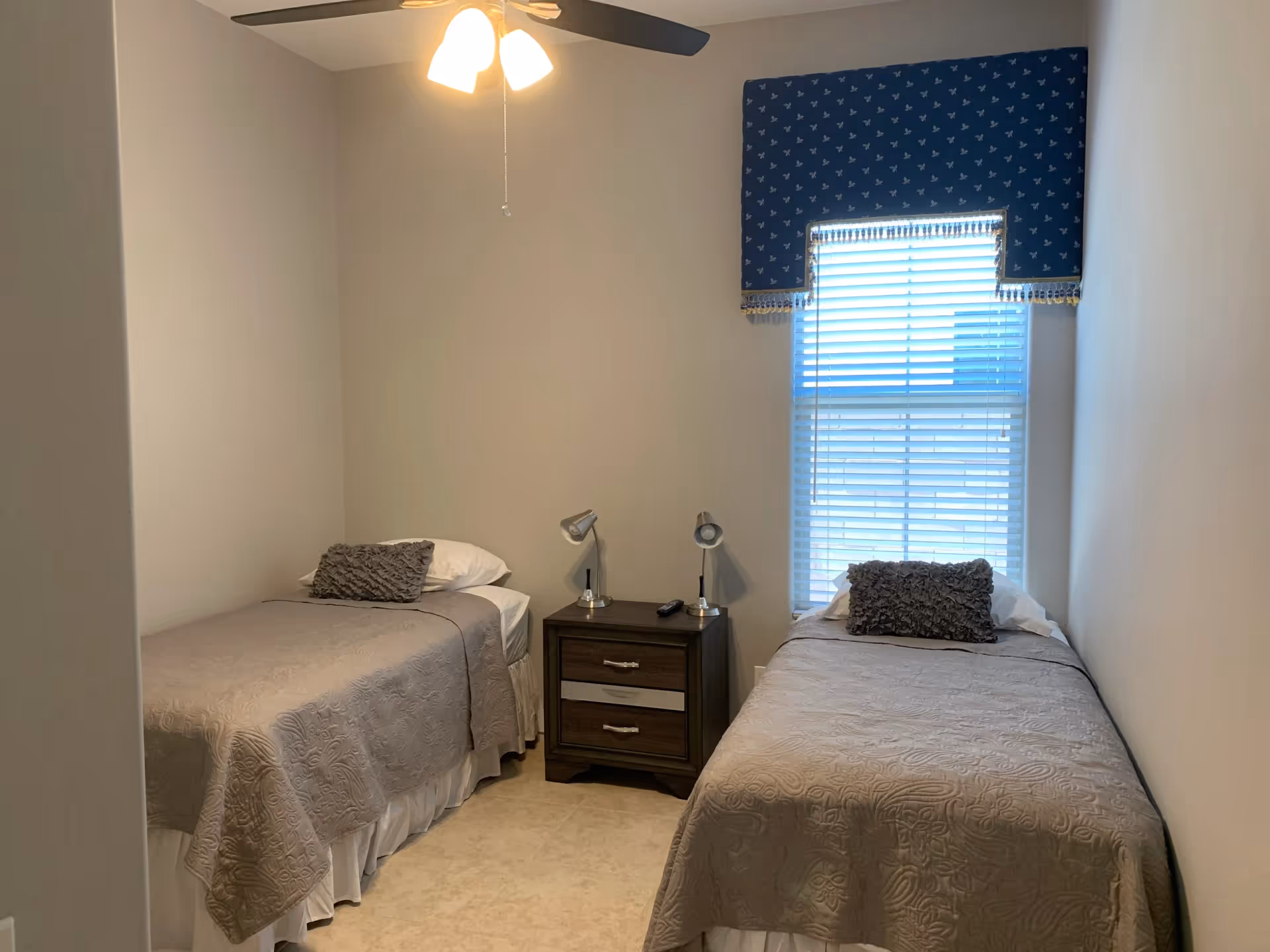 Two twin beds with a nightstand between, ceiling fan, and window with blue valance.