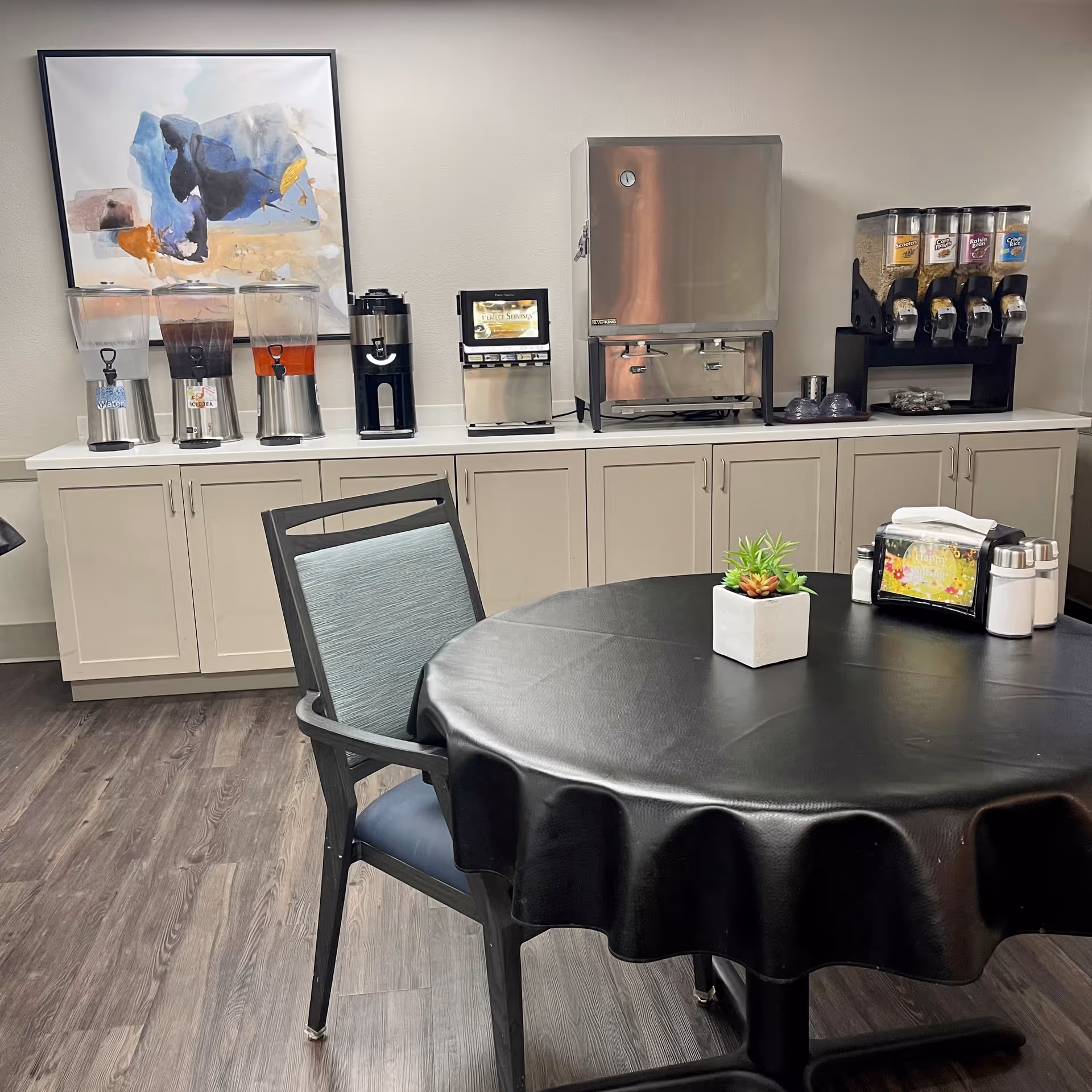 A dining area with a round table covered with a black tablecloth and a chair with a blue cushion. On the table is a small potted plant and a napkin holder with salt and pepper shakers. Behind the table is a counter with beverage dispensers including water, iced tea, and juice, a coffee machine, a hot water dispenser, and cereal dispensers. A colorful abstract painting hangs on the wall above the counter.